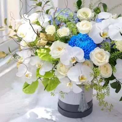 White orchids, roses, and blue chrysanthemum in a grey and white floral box.