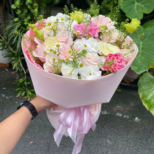 A hand holds a bouquet of pink and white roses, carnations, and daisies wrapped in light pink paper, tied with a pink ribbon.