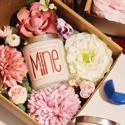 Candle labeled "Mine" surrounded by pink and white flowers in a gift box.