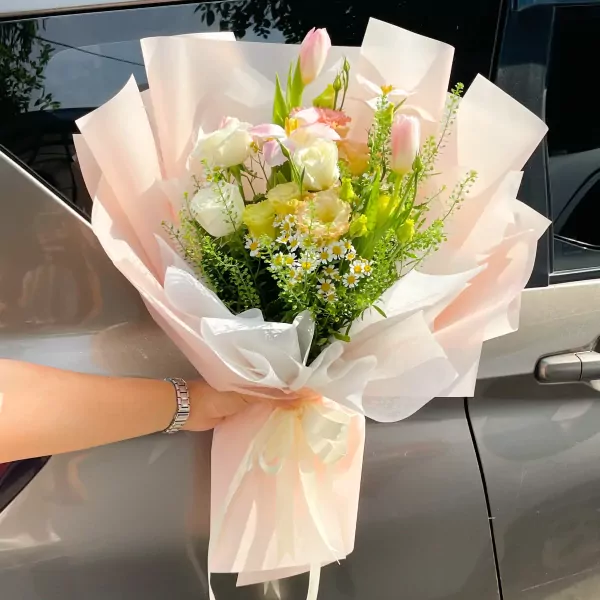 A hand holding a bouquet of pink tulips, white roses, and small white flowers wrapped in pink and white paper.