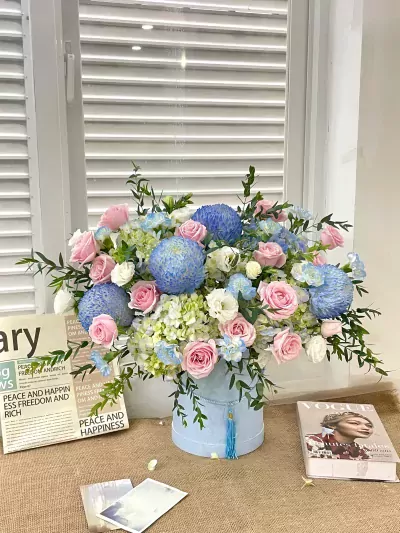 Light blue and pink floral arrangement in a cylindrical box, with books and cards on a burlap surface.
