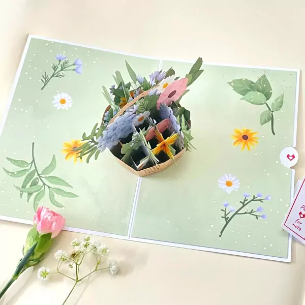 3D pop-up card with a basket of colorful flowers on a light green background with scattered dots and floral illustrations.