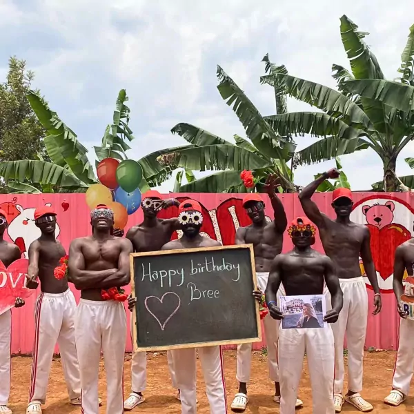 Men with black body paint and red hats hold a chalkboard that says "Happy birthday Bree" and a photo.