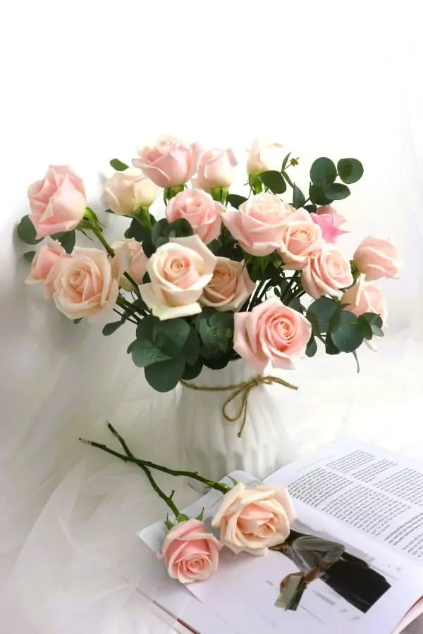 Bouquet of pink roses with eucalyptus leaves in a white vase, two roses on an open book.