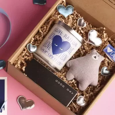 Gift box with candle, plush bear, letter board, and heart embellishments on shredded paper.