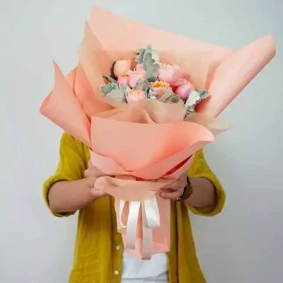 A person in a yellow cardigan holds a bouquet of pink roses, pale green leaves, and lavender flowers wrapped in peach paper.