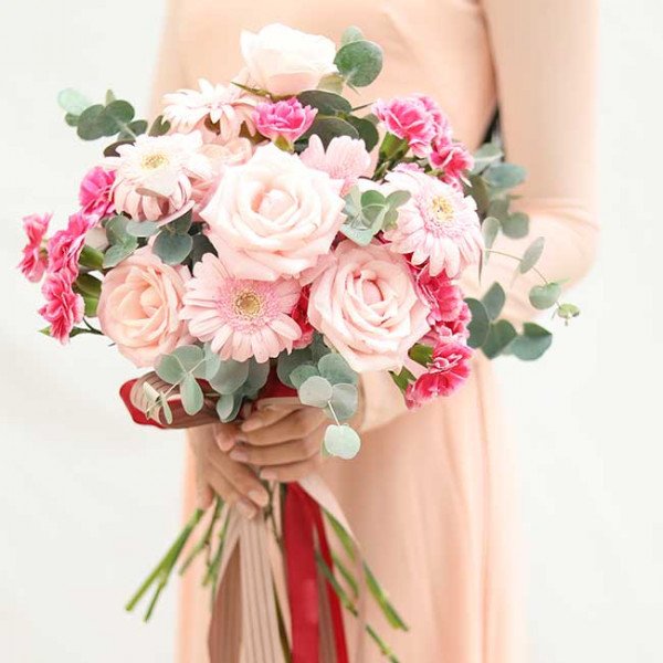 Bridal bouquet with pink roses, gerbera daisies, carnations, and eucalyptus, held by a person in a peach dress.