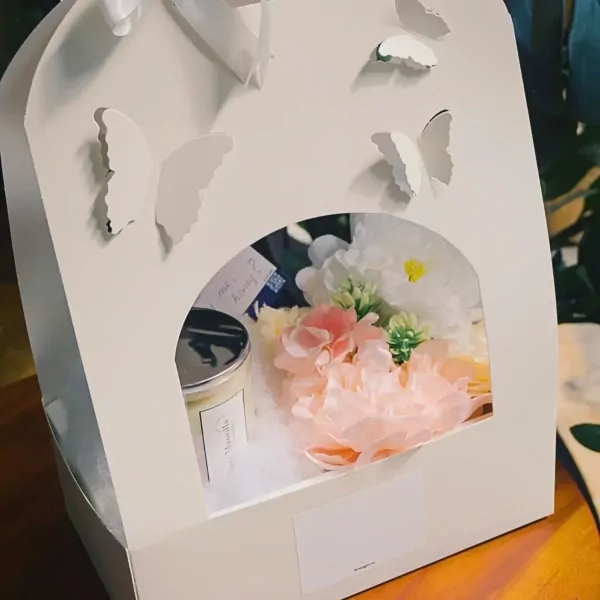 A white gift basket with cut-out butterflies, containing a vanilla scented candle and a bouquet of pink and white flowers.