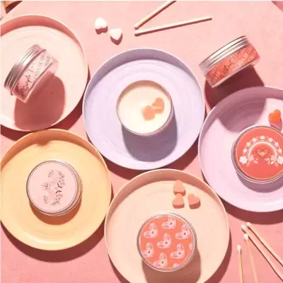 Five pink and peach scented candles in tins arranged on decorative plates, with heart-shaped candies and matches.