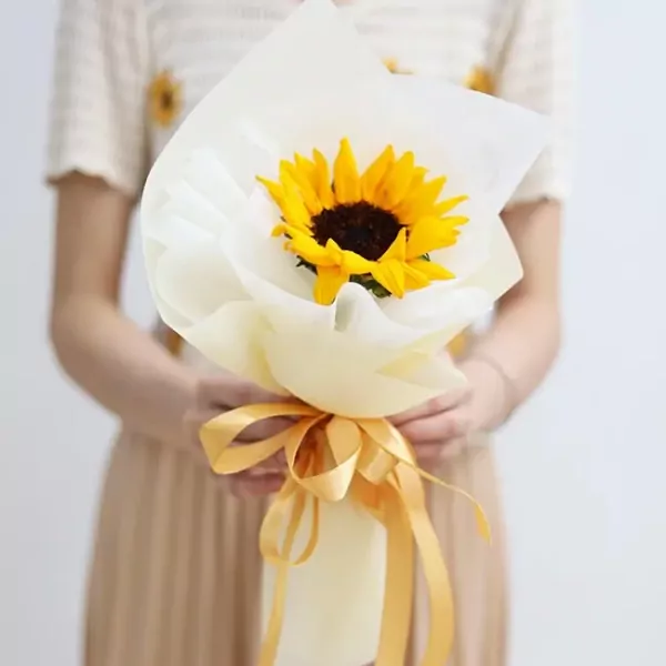 A person holds a bouquet with one sunflower wrapped in white paper and tied with a yellow ribbon.