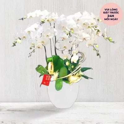 White orchids in a white vase with decorative accents and a red tag.
