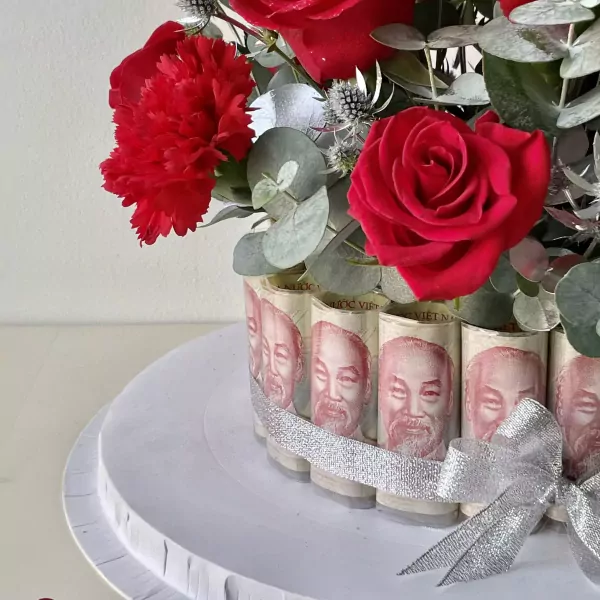 Red roses, carnations, and eucalyptus surround a cake decorated with rolled Vietnamese banknotes and a silver ribbon.