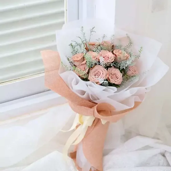 A bouquet of delicate pink cappuccino roses, wrapped in white and peach paper with a pale yellow ribbon.