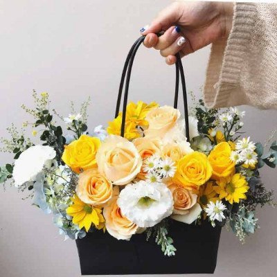 Black basket overflowing with yellow roses, white lisianthus, and assorted greenery.
