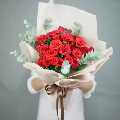 A bouquet of red roses with eucalyptus, wrapped in cream paper and tied with a brown ribbon.