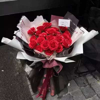 A bouquet of red roses with pearls, wrapped in decorative paper and ribbons, with a "Happy Day" card.