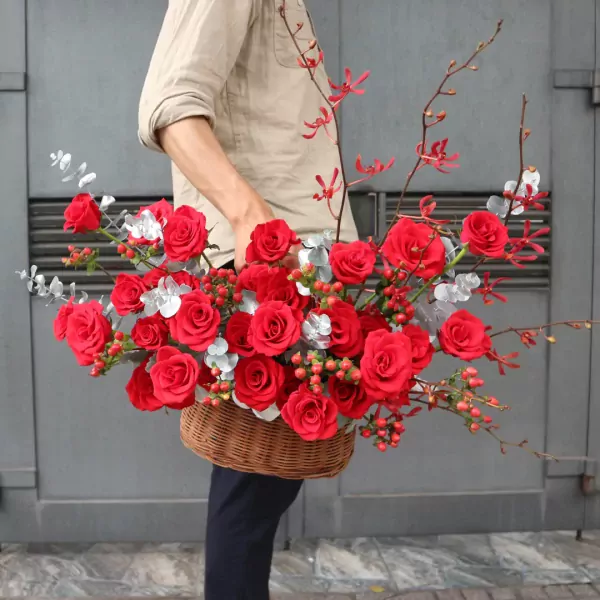 A basket of red roses with delicate red and silver accents held by a person in a tan shirt.
