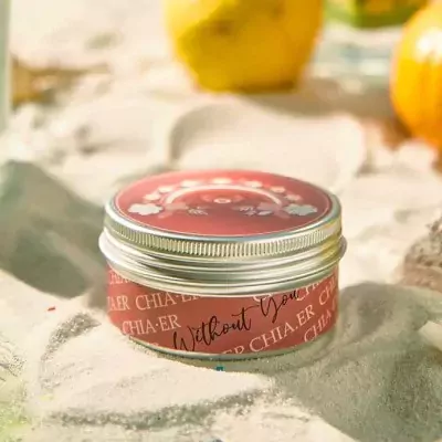 Red scented candle tin with "Without You" text and floral design, surrounded by sand.