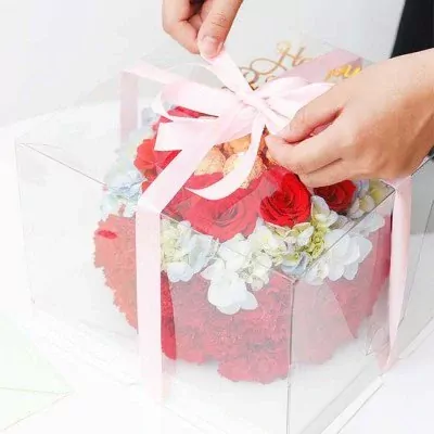 Hands tying a pink ribbon on a clear cake box filled with red roses, hydrangeas, and carnations.