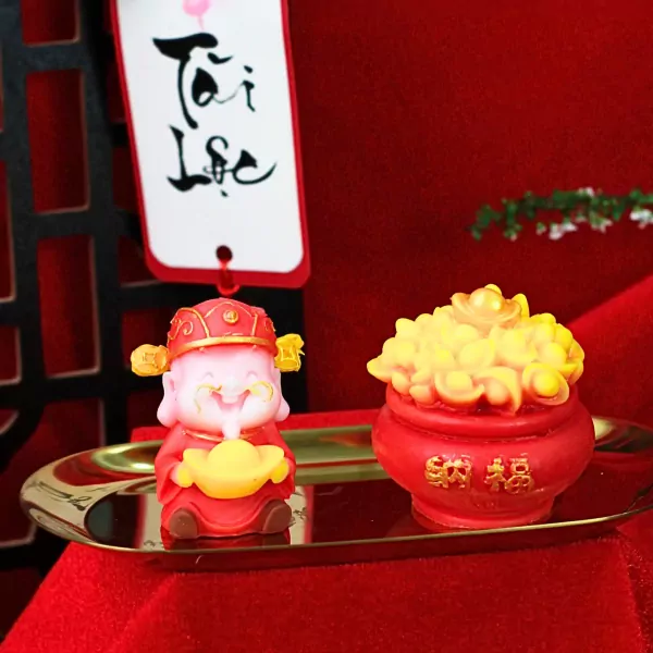 Red wax god holding gold ingot next to red pot of gold coins, on a golden tray.