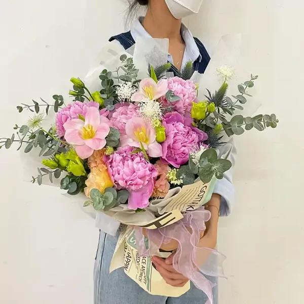 A woman in a blue shirt holds a bouquet of pink peonies, tulips, and carnations with eucalyptus leaves.