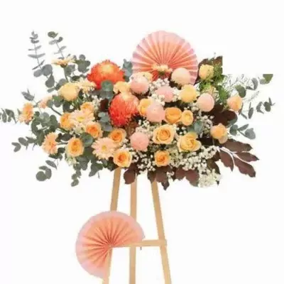 Floral arrangement with orange chrysanthemums, yellow roses, and pink accents on a wooden stand, adorned with paper fans.