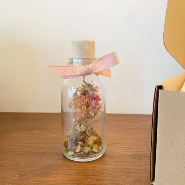 Glass bottle with dried flowers and a pink ribbon on cork stopper, on a wooden surface.