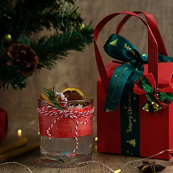 Christmas candle with dried orange, cinnamon, and rosemary in a glass, tied with red and white string, next to a red gift box.