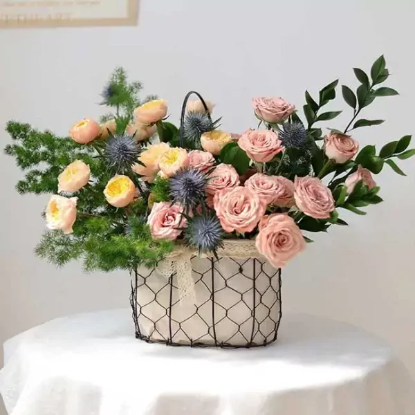 Pink and peach roses, blue thistles, and greenery in a wire basket.