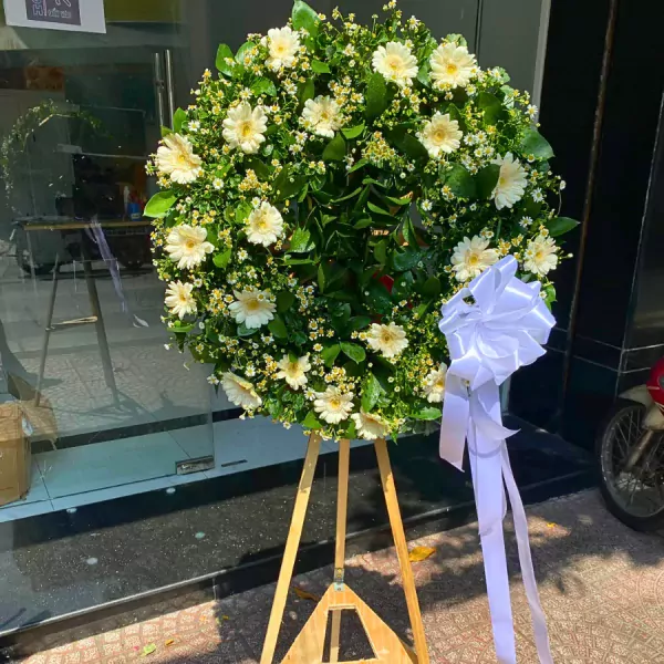 White gerbera daisies and baby's breath wreath on a wooden easel with a white ribbon.