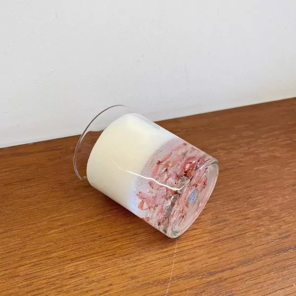 A clear glass candle with white wax and pink petals embedded in the bottom half.