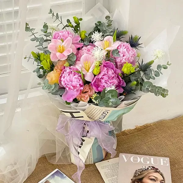 A bouquet of pink peonies, tulips, and lisianthus with eucalyptus and thistles, wrapped in newspaper and sheer fabric, tied with a purple ribbon.