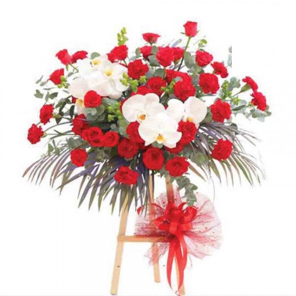 Floral arrangement with red roses, white orchids, and greenery on a wooden stand with a red bow.