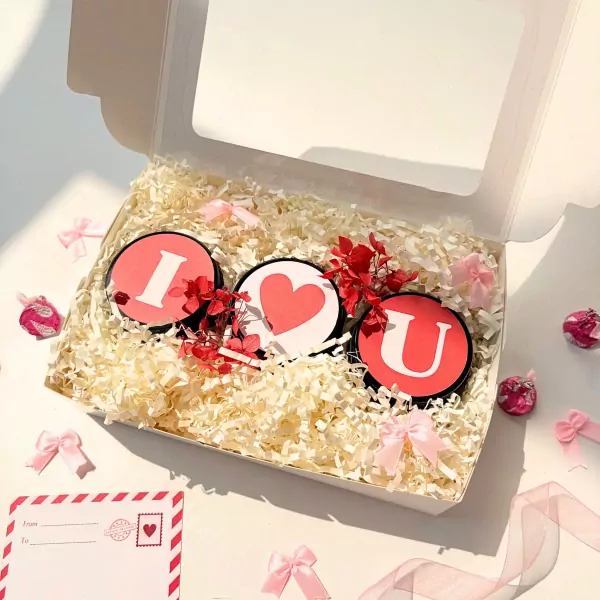 Gift set with three round candles spelling "I Heart U" nestled in shredded paper, surrounded by pink bows and dried flowers.