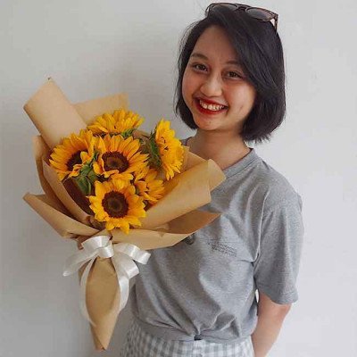 Smiling woman holding a bouquet of vibrant sunflowers wrapped in brown paper with a white ribbon.