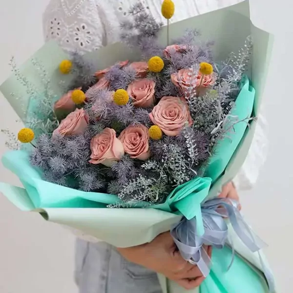 A bouquet of blush pink roses, craspedia, and delicate grey foliage wrapped in mint green paper, tied with a grey ribbon.