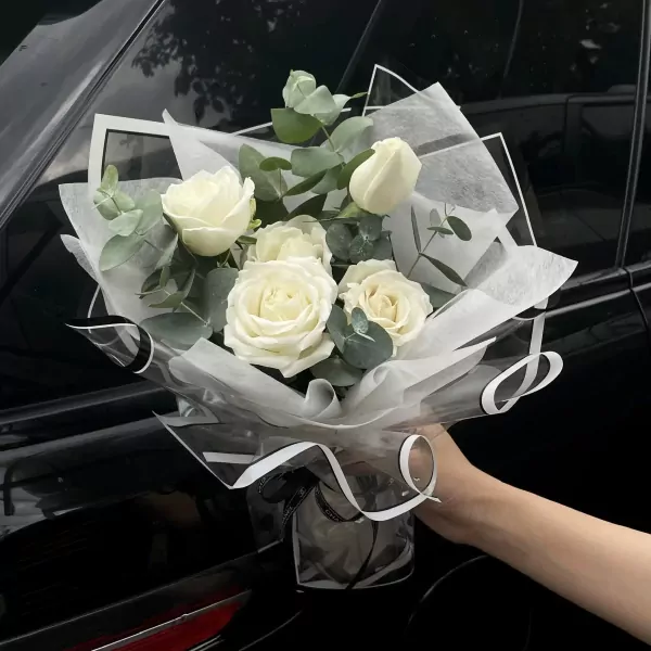 Bouquet of white roses with eucalyptus, wrapped in sheer white paper and black ribbon, held by a hand.