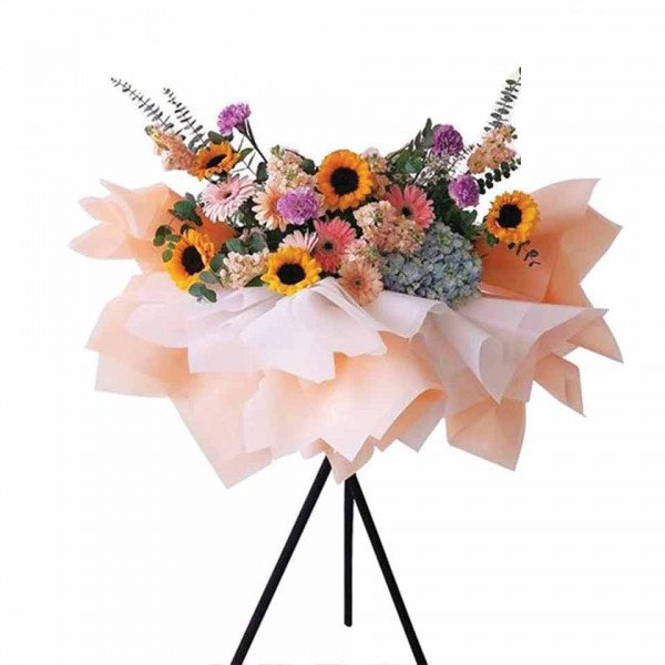A large, colorful flower arrangement with sunflowers, hydrangeas, and gerbera daisies, presented on a stand.
