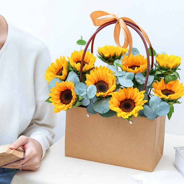 Brown paper gift bag filled with sunflowers and eucalyptus, tied with an orange ribbon.