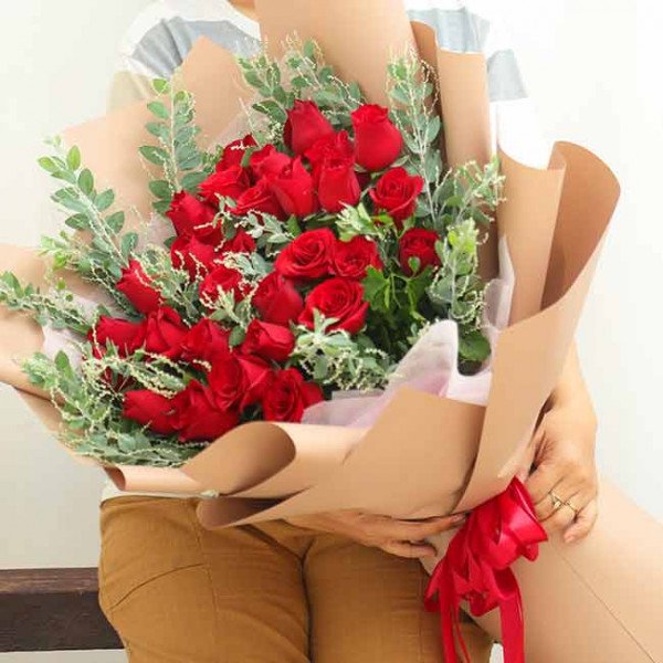 A bouquet of red roses with greenery, wrapped in brown paper and tied with a red ribbon.