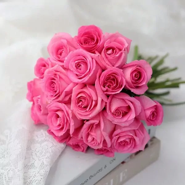 A bouquet of 20 bright pink roses with water droplets, resting on a lace cloth and a book.