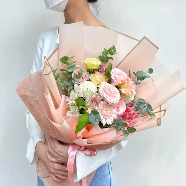A person in a white shirt and blue jeans holds a bouquet of pink, white, yellow, and orange flowers wrapped in pink paper.