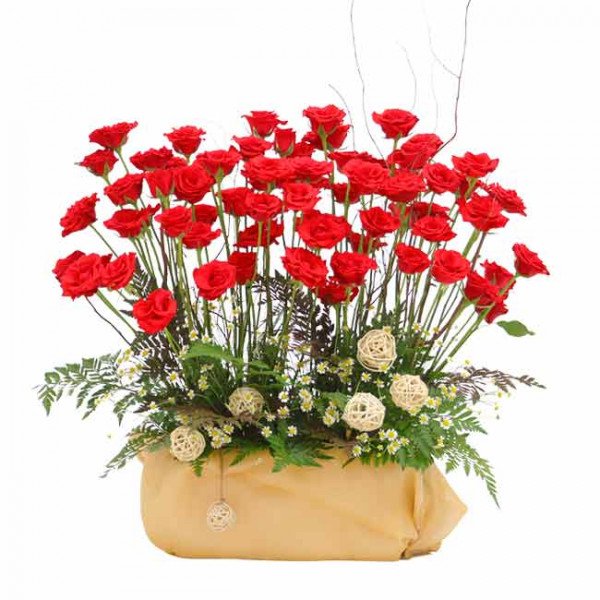 A large bouquet of red roses in a tan rectangular vase with decorative filler flowers and woven balls.