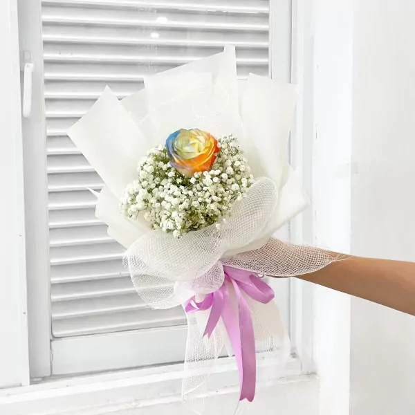 Rainbow rose bouquet with baby's breath, wrapped in white paper and tied with a purple ribbon.