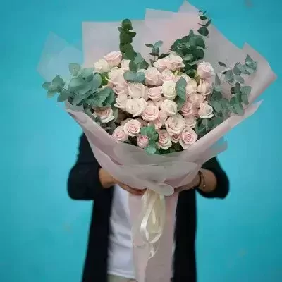 Bouquet of pink and white roses with eucalyptus leaves, wrapped in pink paper, held by a person.