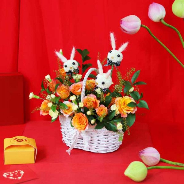 A white basket overflowing with orange and yellow roses, decorated with bunny figures.