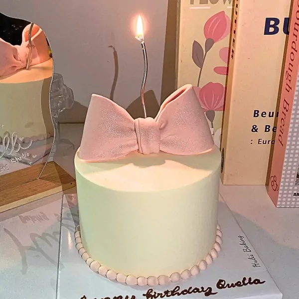 A pale yellow cake topped with a pink bow and a lit candle, adorned with tiny pearls at the base.