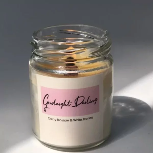 A lit candle in a glass jar with a label that reads "Goodnight Darling, Cherry Blossom & White Jasmine."