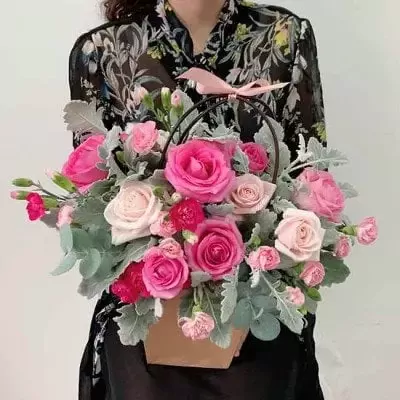 A basket of pink and red roses with greenery, held by a person in a floral top.