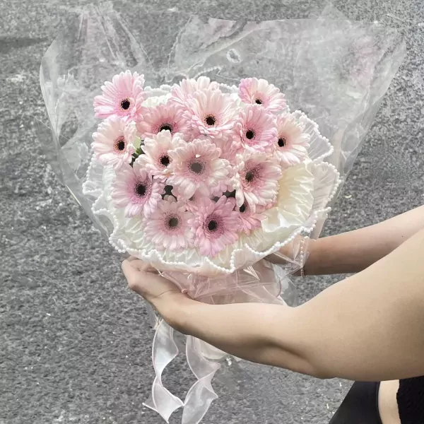 Bouquet of pink gerbera daisies wrapped in clear cellophane and white frills with pearl accents.